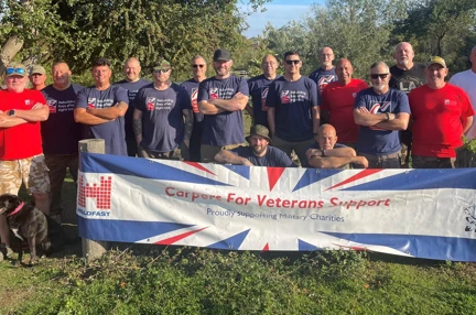 A group photo of all the competitors from this years competition. The group are stood outside on the grass with trees behind them and in front of them is a large banner that reads 'Carpers for veterans support'