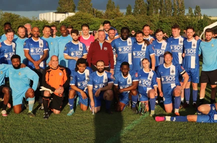 A group shot containing both teams in their football kit and match officials on the pitch. Half the group are kneeling at the front and the others are standing behind with arms around one another's shoulders