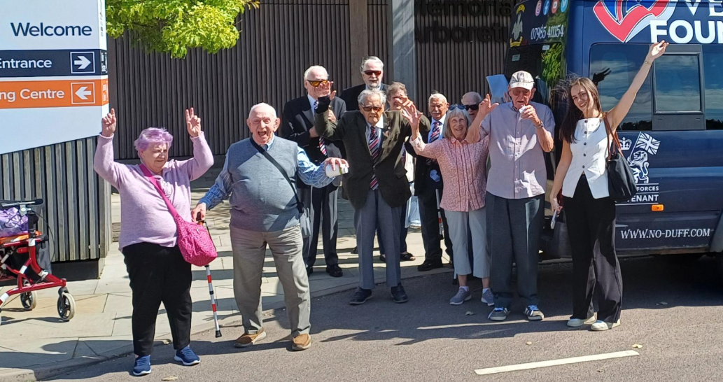 A group photograph of blind veterans lifting their hands in the air as they stand next to a welcome sign for the National Memorial Arboretum