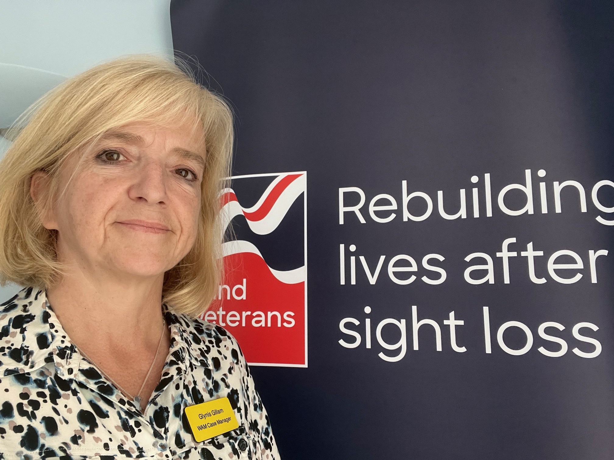 Glynis smiles and stands in front of a Blind Veterans UK branded banner