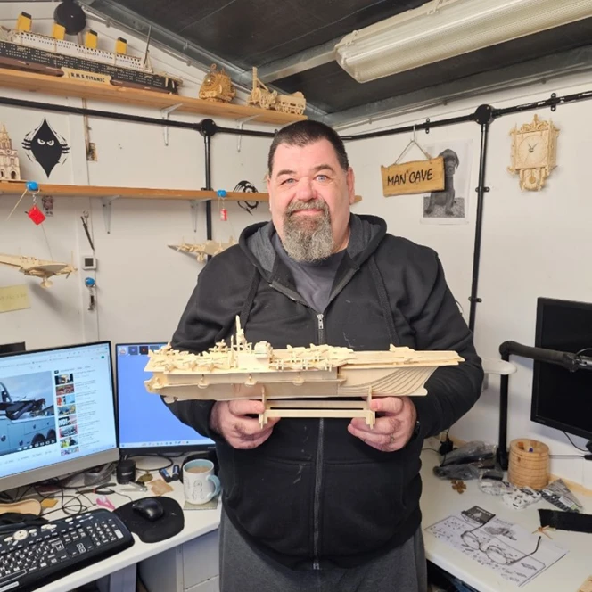 Gary is proudly holding a meticulously crafted wooden model of a ship, as he stands in his personal workshop, a space warmly labelled with a sign on the wall that reads "Man Cave".
