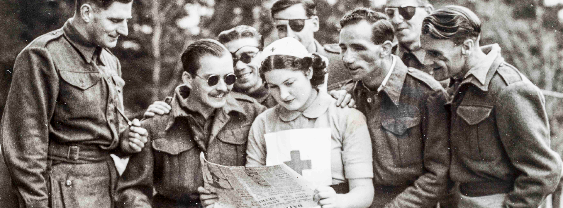 A nurse standing while reading a newspaper, as a group of blind veterans are gathered round her 