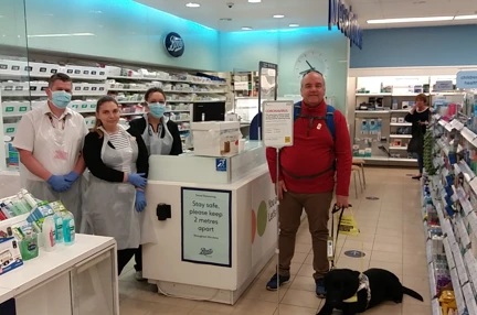 John is being served in a pharmacy with his guide dog by his feet