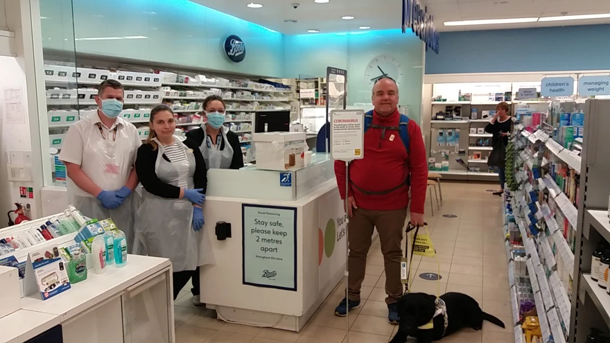 John is being served in a pharmacy with his guide dog by his feet