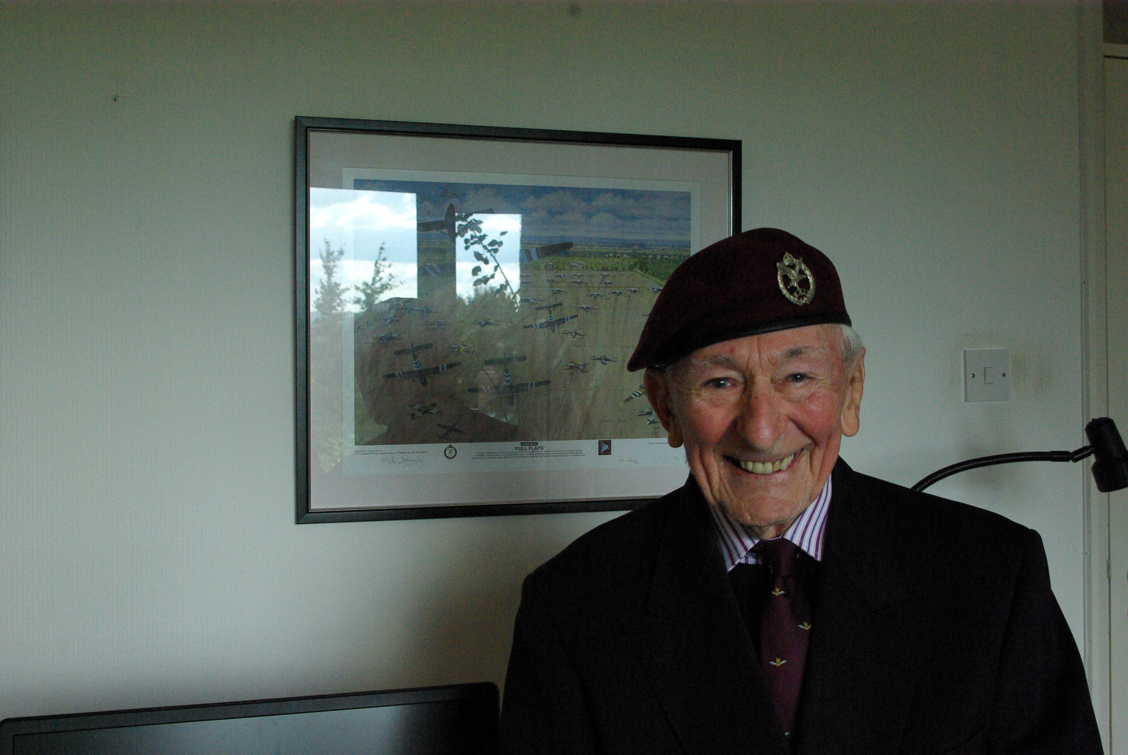 Blind veteran Jim smiling as he stands in front of a framed piece of artwork showing the Horsa Gilders landing at Arnhem