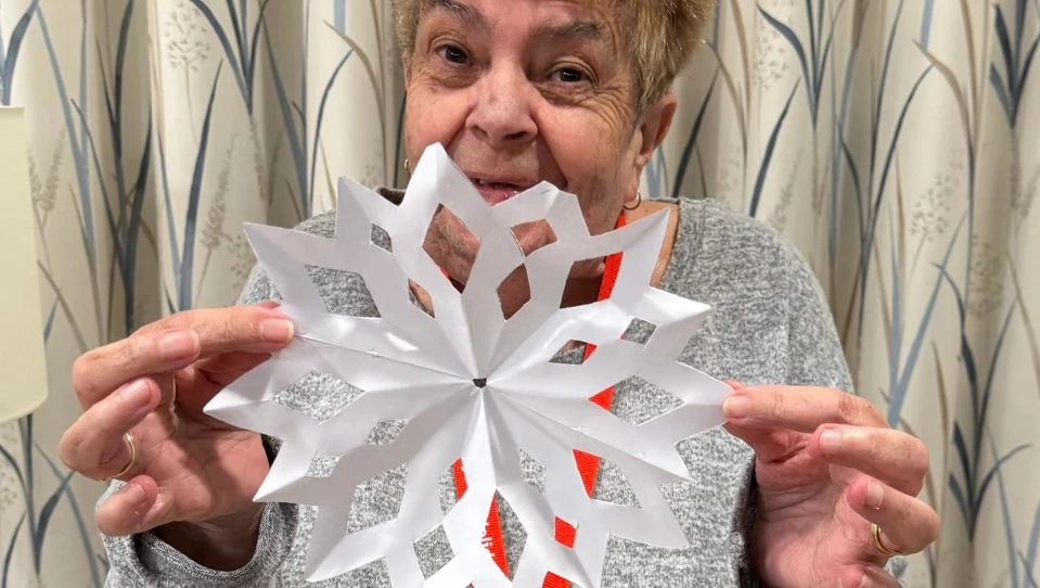 he image shows Maureen, wife of blind veteran Jeff, smiling warmly as she holds up a beautifully crafted paper snowflake during a visit to the Rustington Centre.
