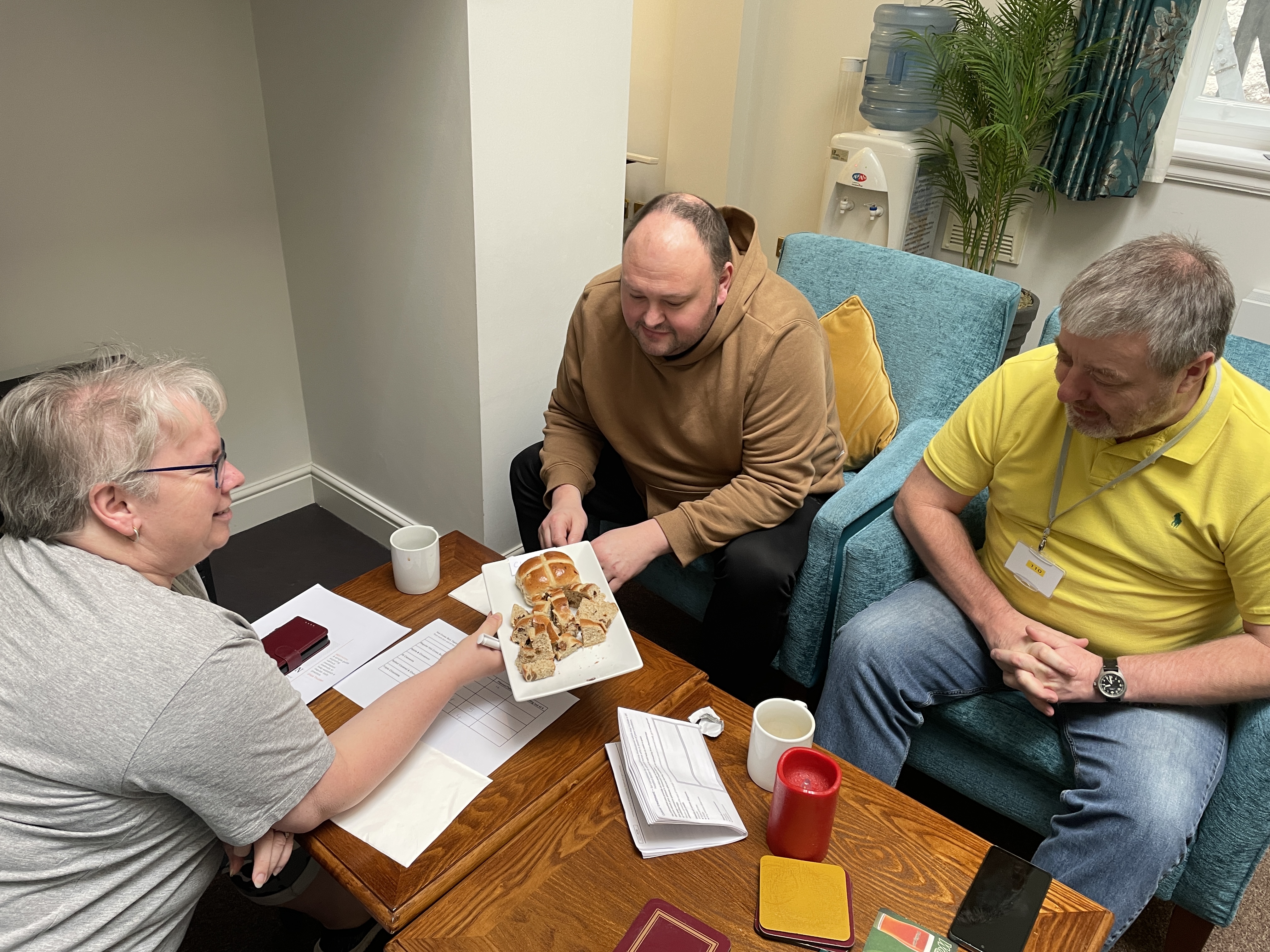 Matt and Ivan sit opposite Trudy who is holding out a plate of hot cross buns to them.