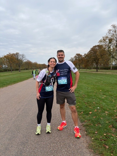 Ed and Nicola are dressed in their Blind Veterans UK running t-shirts during a run. They have their arms around each other and have stopped for a photo along a wide tarmac path.