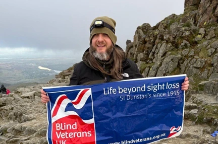 Benjamin at the top of Snowdon holding a Blind Veterans UK banner. Benjamin is wearing a beanie hat and a headtorch.