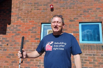 Lee holding is cane and wearing a Blind Veterans UK top is standing outside the swimming pool where he will complete his challenge smiling