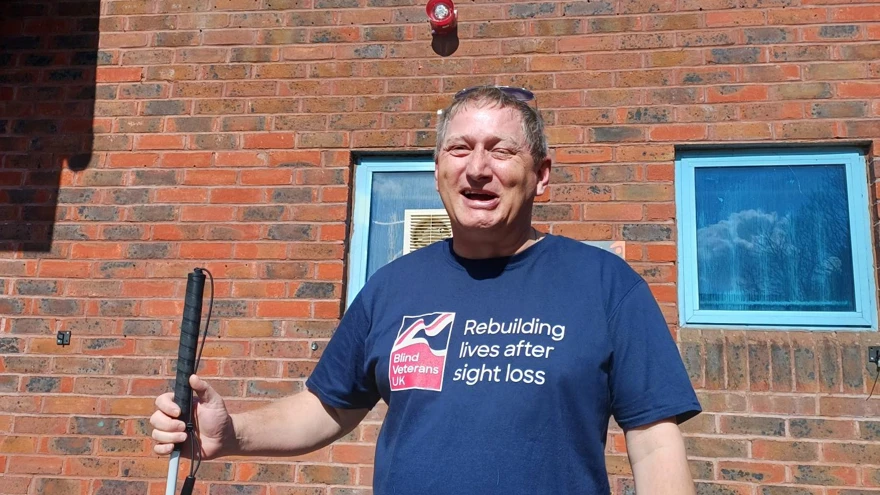 Lee holding is cane and wearing a Blind Veterans UK top is standing outside the swimming pool where he will complete his challenge smiling