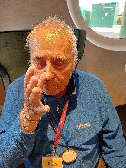 A man in a blue top sits with his eyes closed and his right hand raised to his nose pressing his right nostril closed.