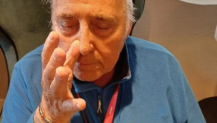 A man in a blue top sits with his eyes closed and his right hand raised to his nose pressing his right nostril closed.