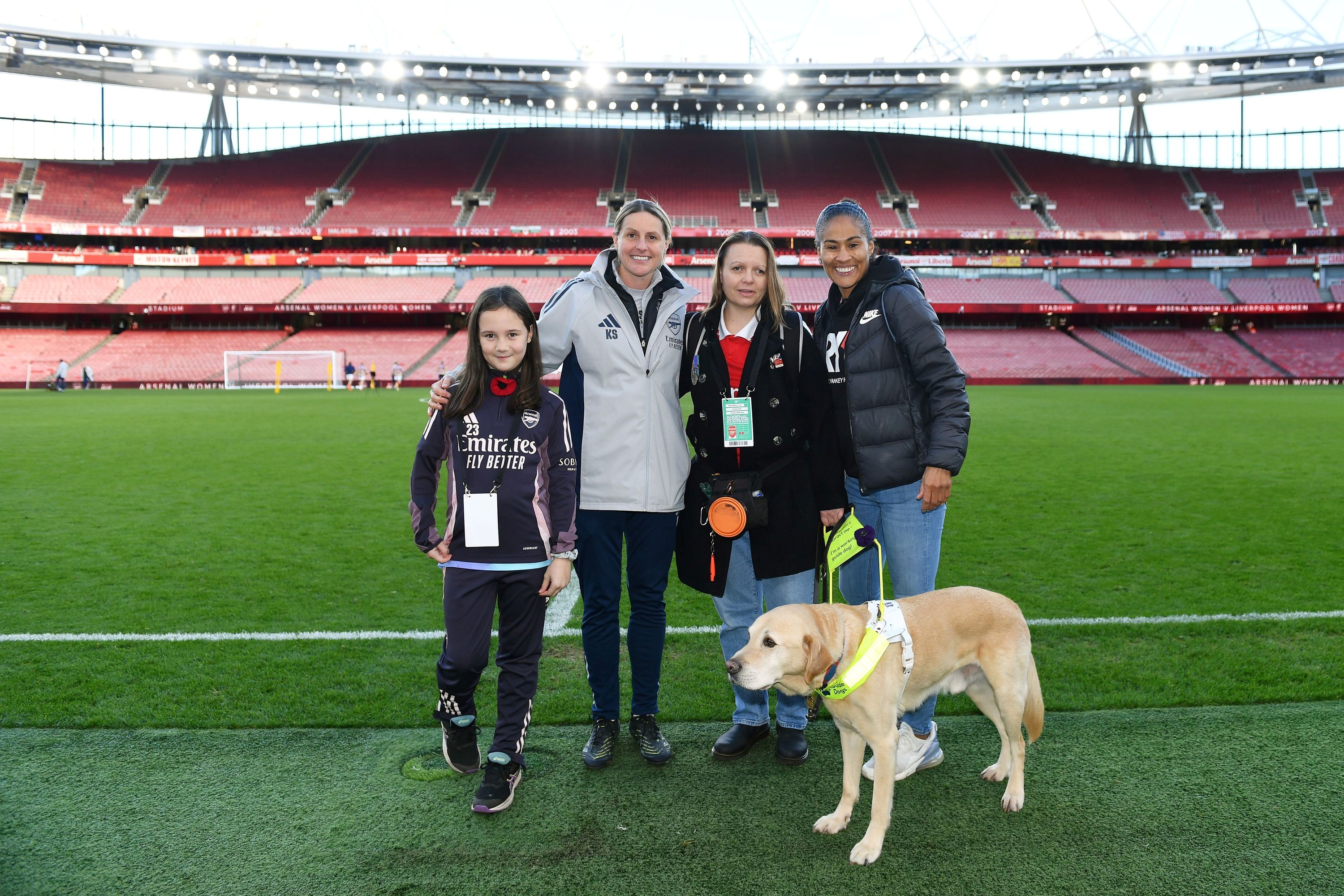 Kelly and Archie stand between Kelly's heroes, footballers Rachel Yankey and Kelly Smith. Bethany is stood the other side of Kelly Smith. They are stood at the side of the pitch at the Emirates Stadium.