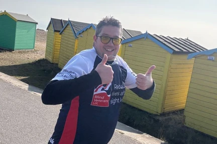 Wayne with both thumbs up looking towards the camera as he runs along the promenade at Rustington. He is wearing a Blind Veterans UK t-shirt and yellow tinted glasses