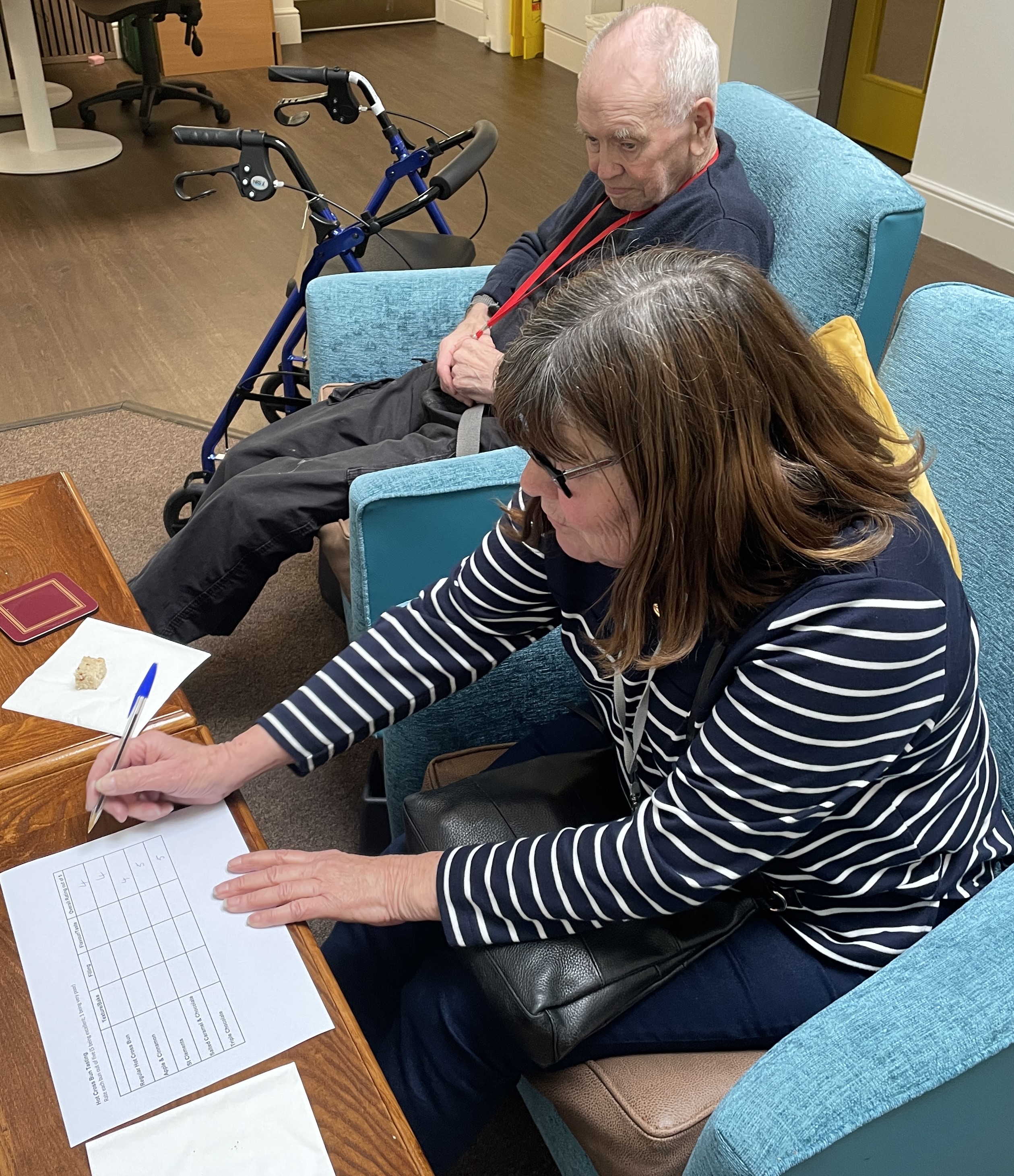 Bob and Sylvia sit side by side in armchairs, Sylvia is filling in scores on a judging form.