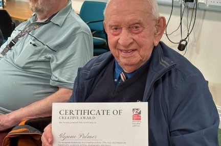 Glynne is seated at a table, smiling as he holds a certificate for creativity.
