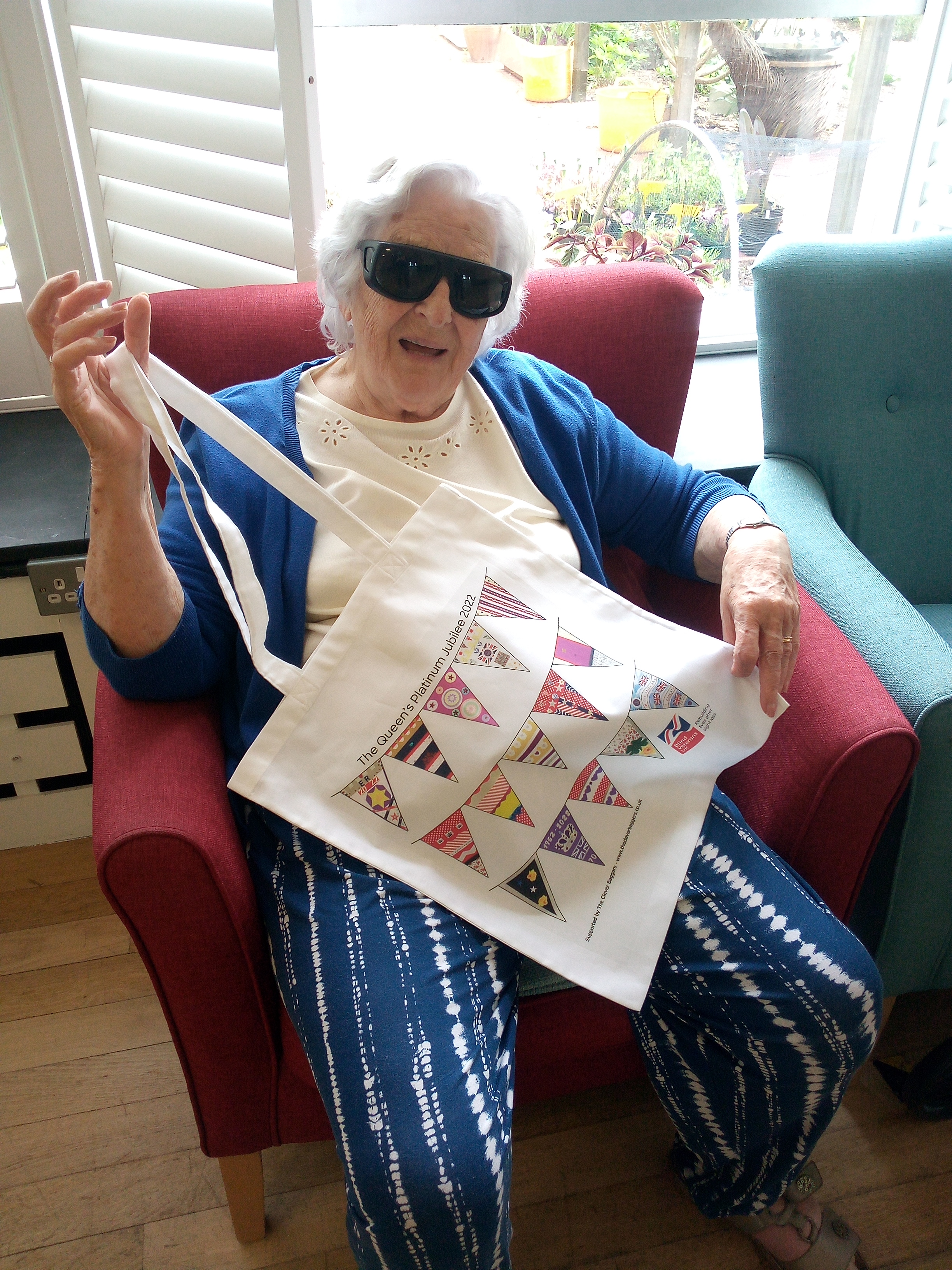 Blind veteran Betty pictured sitting down on a chair holding up her jubilee bunting bag