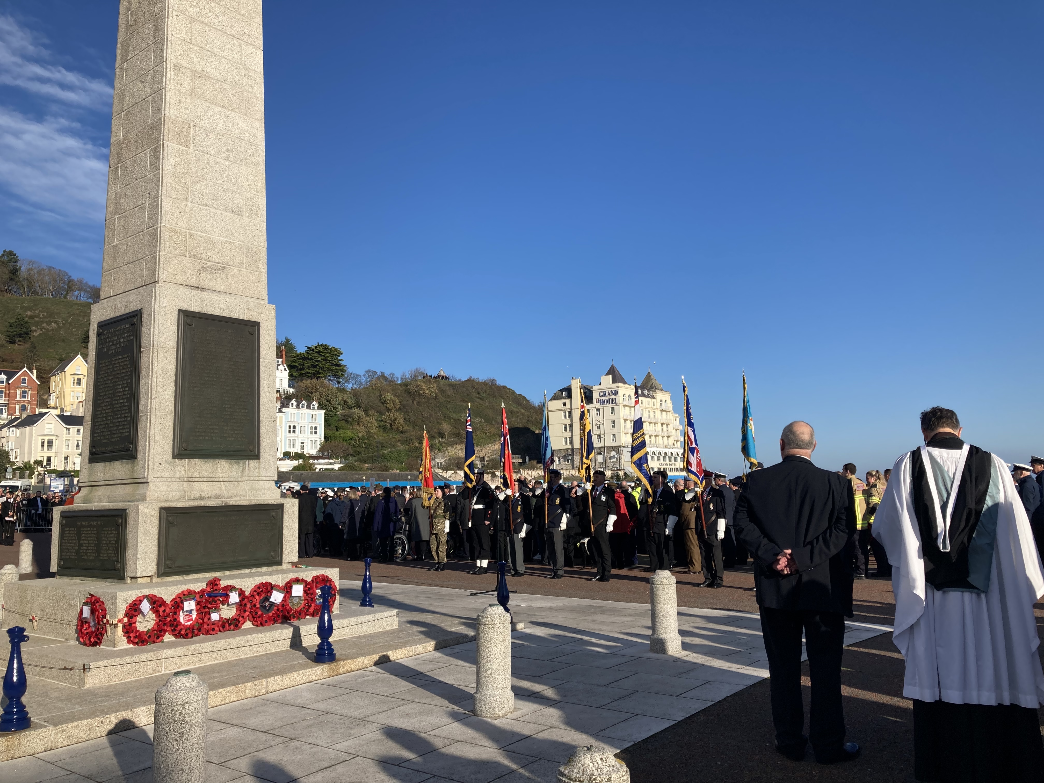 Llandudno town war memorial with poppy wreaths at its base.  Standard bearers and veterans can be seen looking towards the memorial in the background.