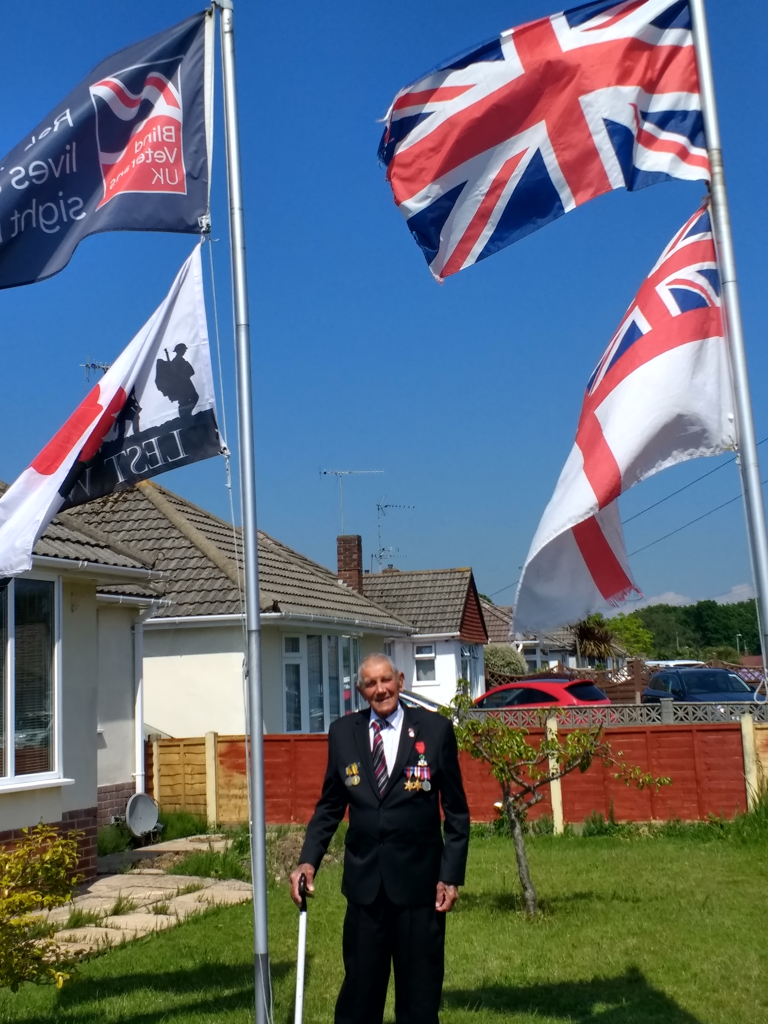 Blind veteran Eddie standing between two flag poles in his garden, displaying a Blind Veterans UK, Union Jack and Lest We Forget flag 