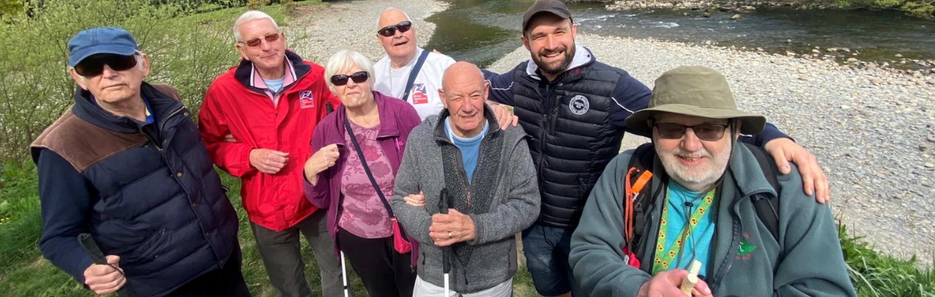 Blind veterans, smiling enjoying a group walk at the Llandudno centre. Some have canes. They are by a river with trees in the background