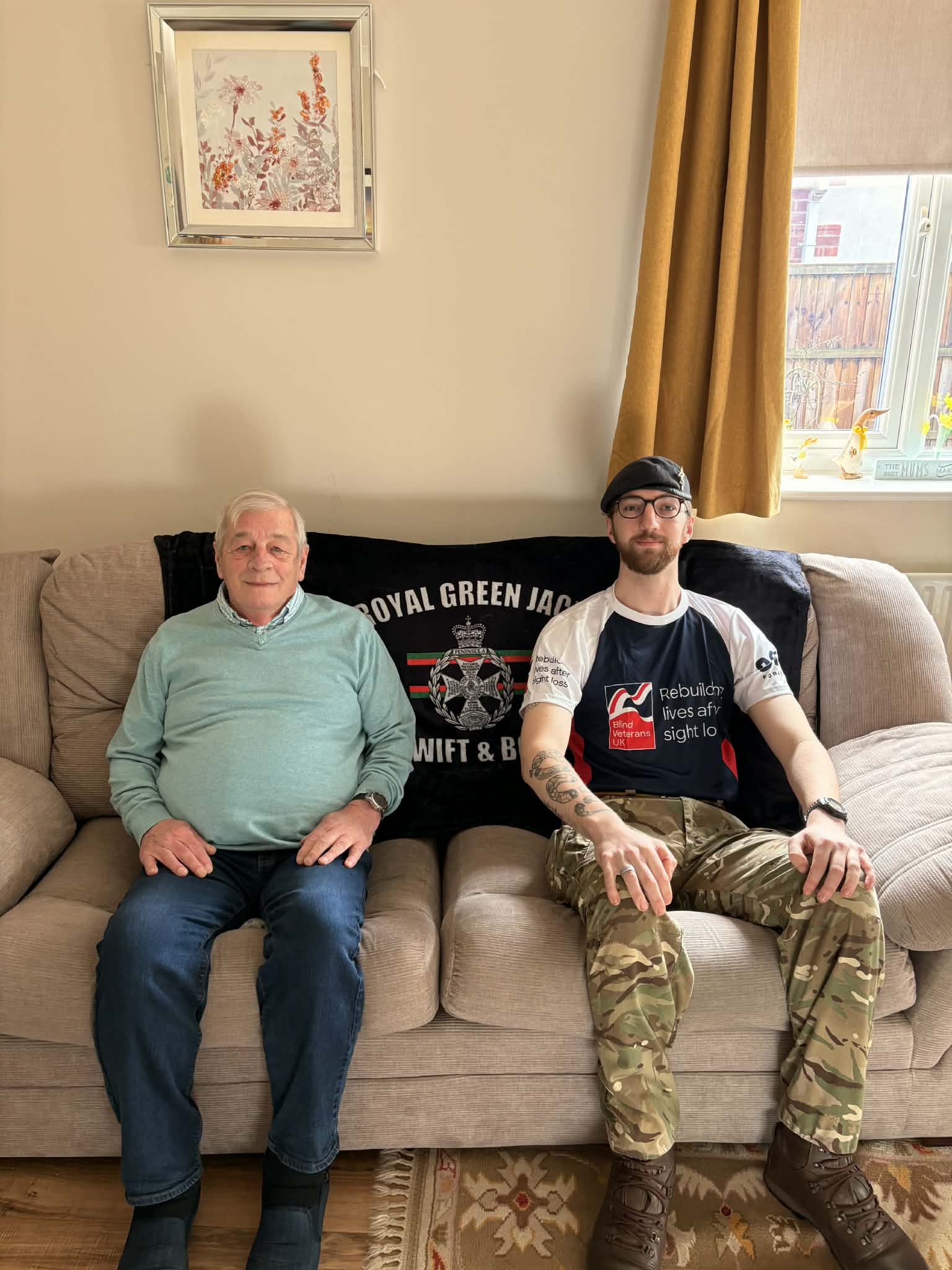 Jack and his grandad are sat next to each other on a sofa with a Royal Green Jackets flagged behind them. Jack is wearing his Army trousers and a Blind Veterans UK t-shirt.