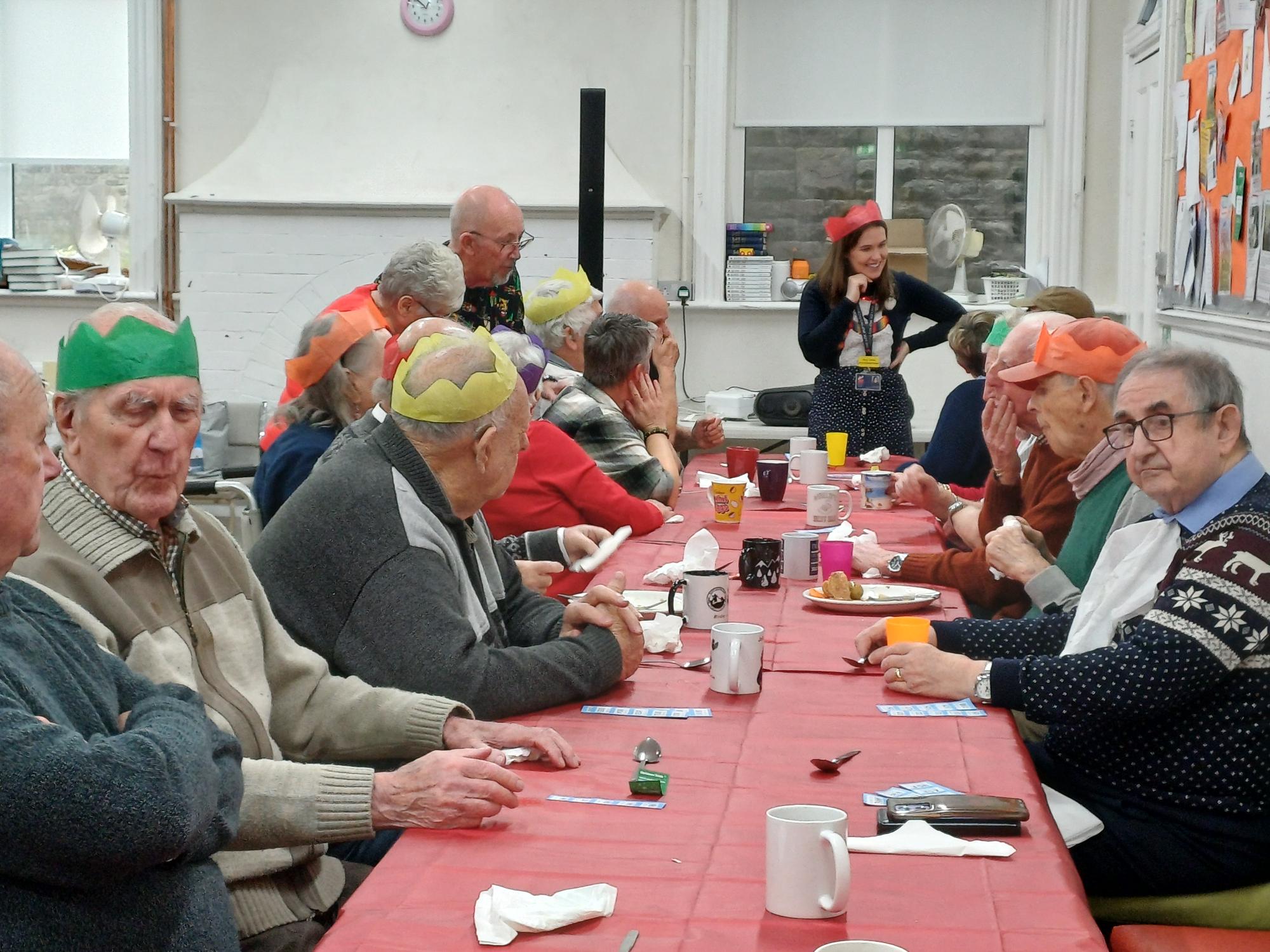 A group of veterans sat along both sides of a long table, many wearing hats from crackers. The group are chatting to one another and enjoying the festivities.