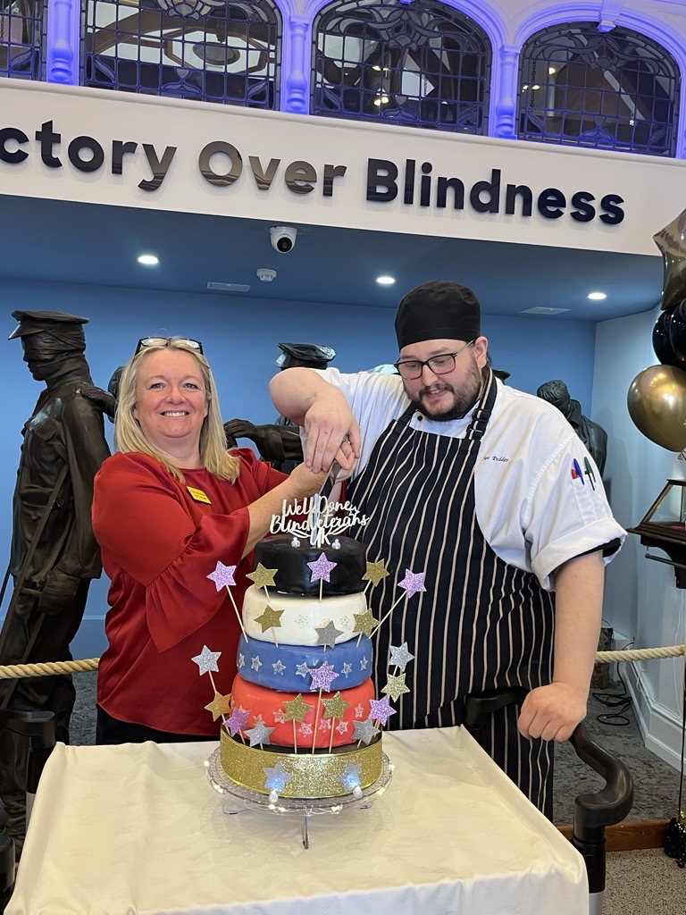 Kathy and Joe stood in front of the Victory over Blindness sculpture at the Llandudno Centre. They are ready to cut the five tier cake covered with stars and a cake topper that reads "Well done Llandudno"