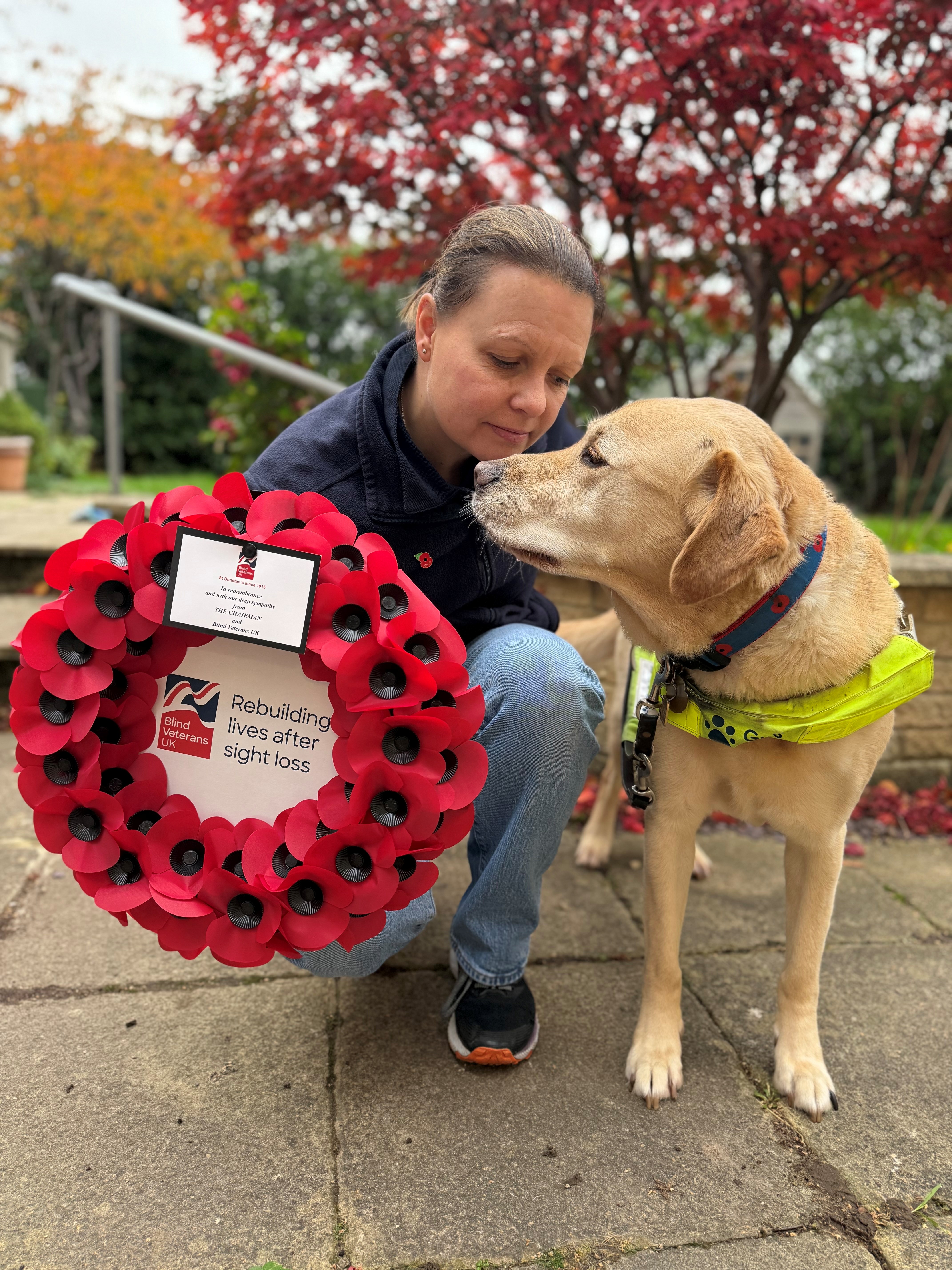 Kelly knelt down next to Archie, a golden retriever cross. The pair are looking into each others eyes. Archie is wearing his Guide Dog harness and a poppy collar. Kelly is holding a wreath.