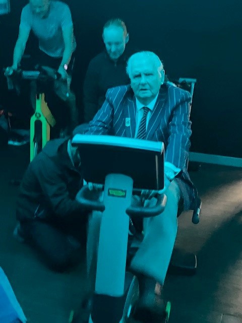 Norman is sat on an exercise bike wearing a striped blazer jacket and tie