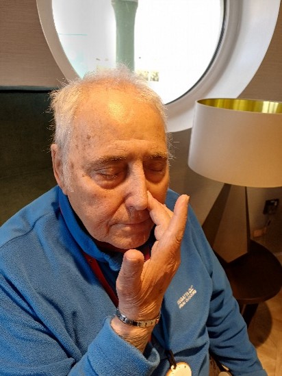 A man in a blue top sits with his eyes closed and his right hand raised to his nose pressing his left nostril closed.