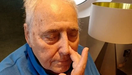 A man in a blue top sits with his eyes closed and his right hand raised to his nose pressing his left nostril closed.