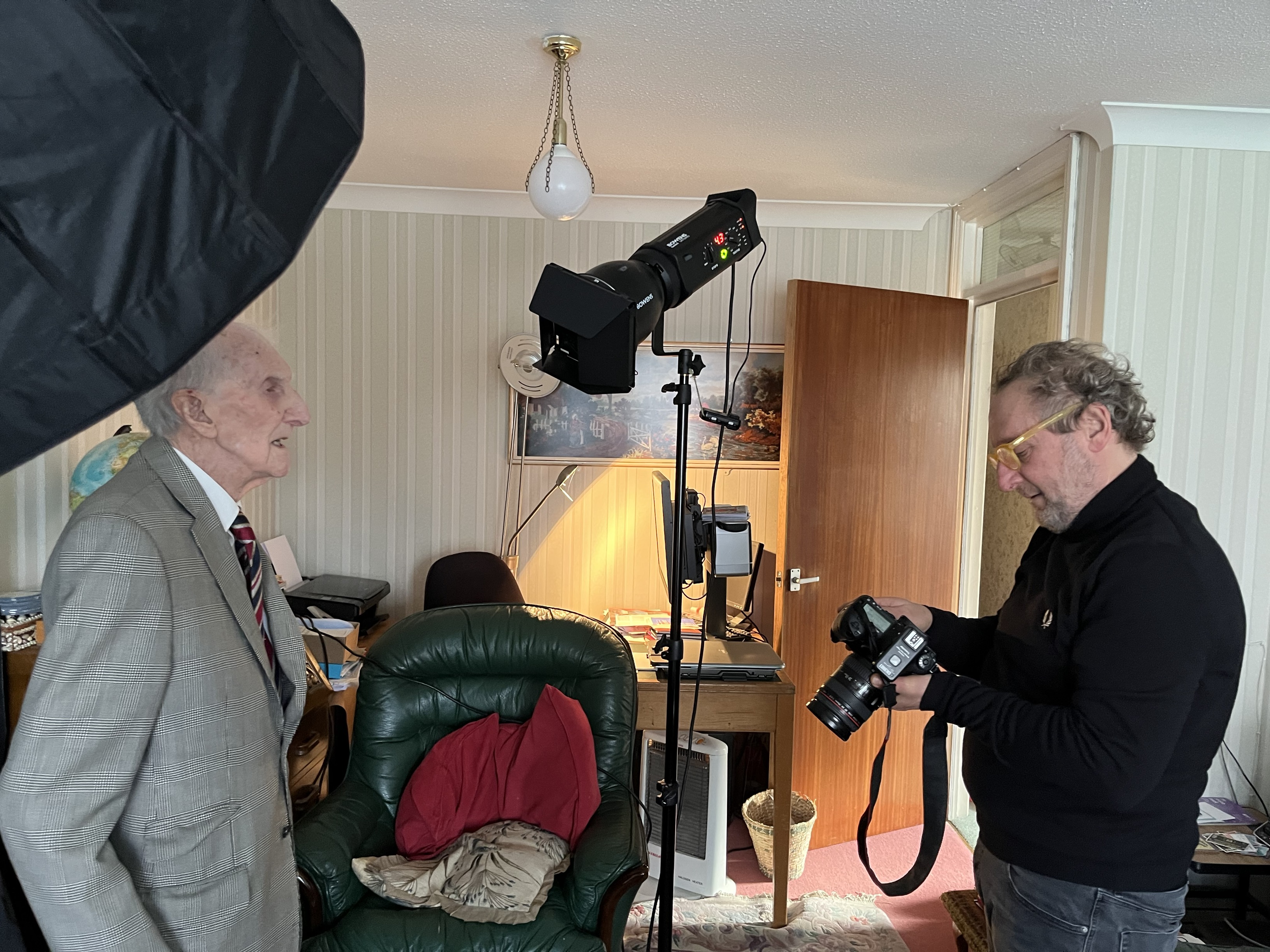 Peter stands for a photograph in his living room, as Richard checks his camera. Lighting equipment and a black backdrop are seen in the background.