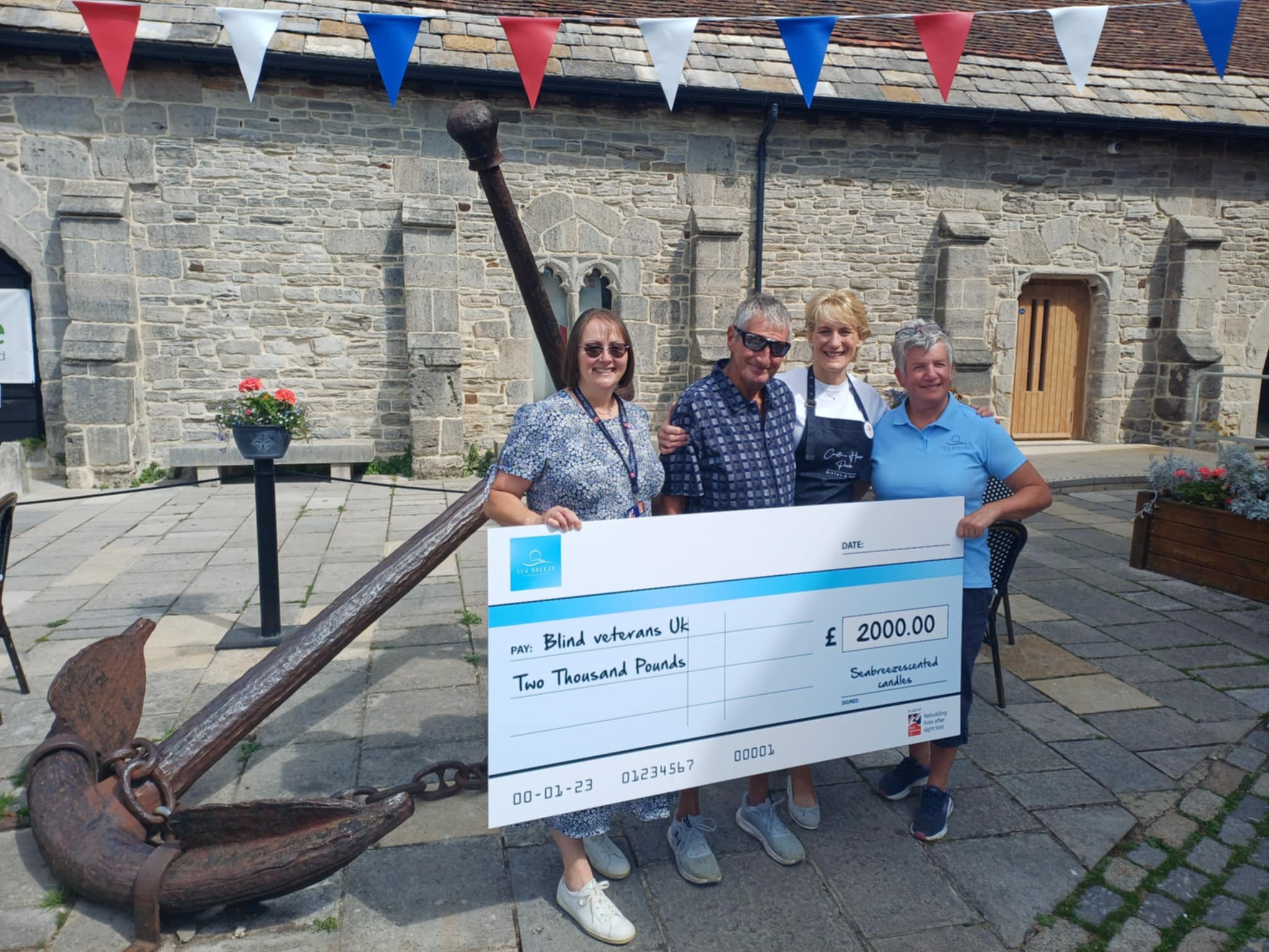 Tony passes an oversized cheque over to a representative from Blind Veterans UK. He is joined by his wife and a bistro owner who helped sell the canldles.