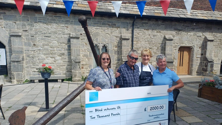 Tony passes an oversized cheque over to a representative from Blind Veterans UK. He is joined by his wife and a bistro owner who helped sell the canldles.