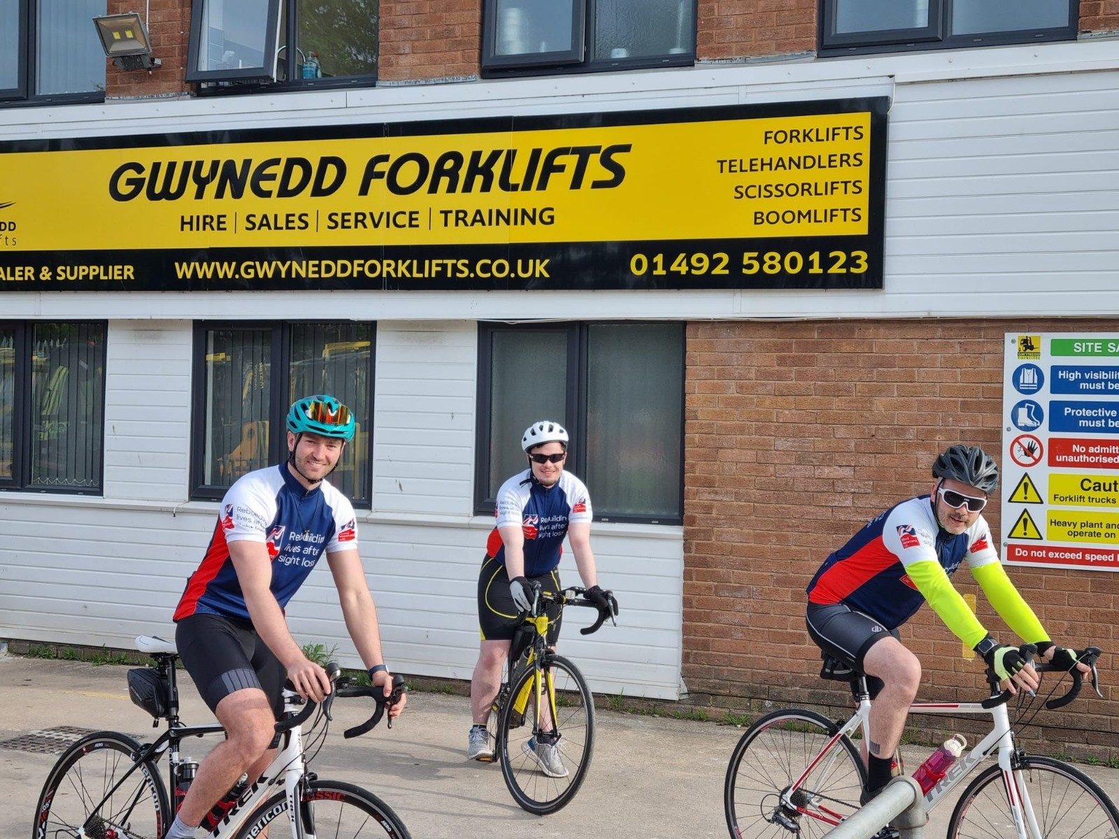 The three cyclists are on their bikes and ready to start their challenge below a large yellow sign for Gwynedd Forklifts