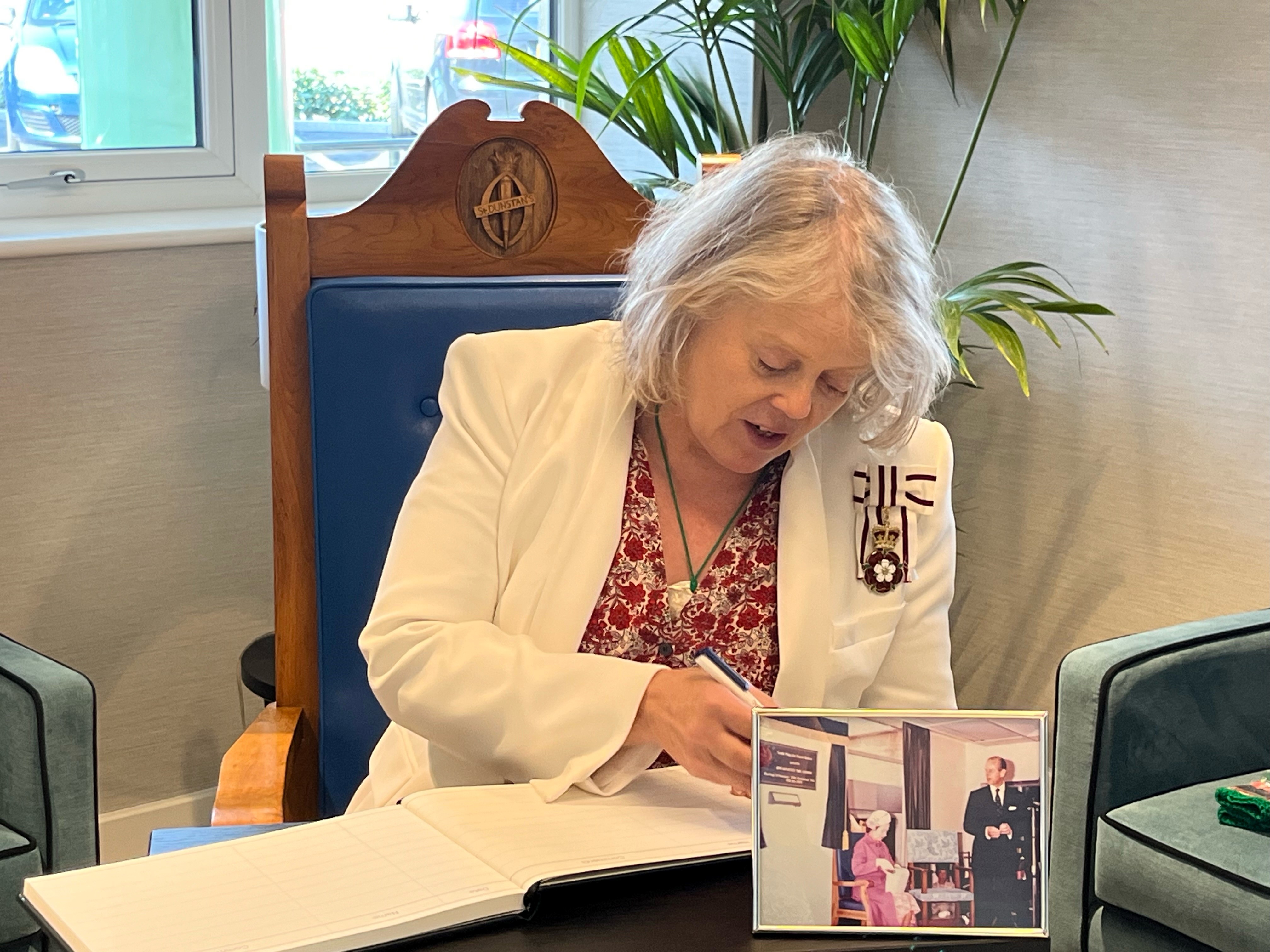 Lady Emma is sat on the Queen's chair with a framed picture of the Queen sat in the chair next to her. Emma is signing the guestbook