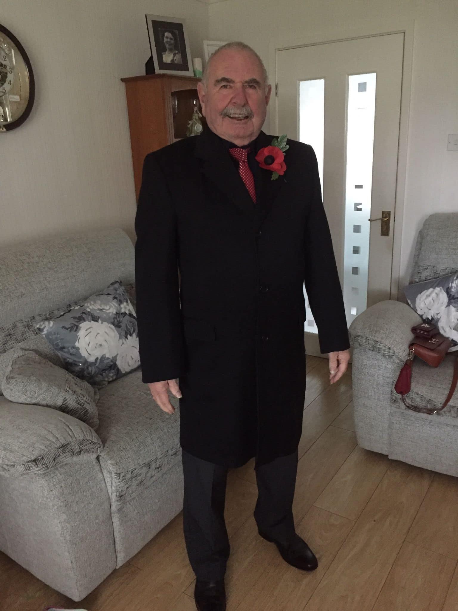 Joe is stood inside a living room wearing a smart black coat, red tie and large poppy
