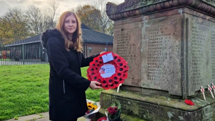 Stephanie stands next to the war memorial with wreaths already laid holding the wreath she is going to place