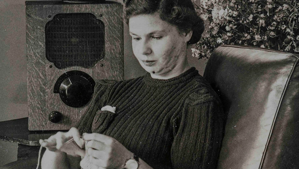 An archive photograph of a young blind veteran Barbara sitting on an armchair and sewing