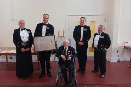 George is sat between four members of the HMS Sultan Mess. The Commanding Officer holds the photograph gifted to them by George.