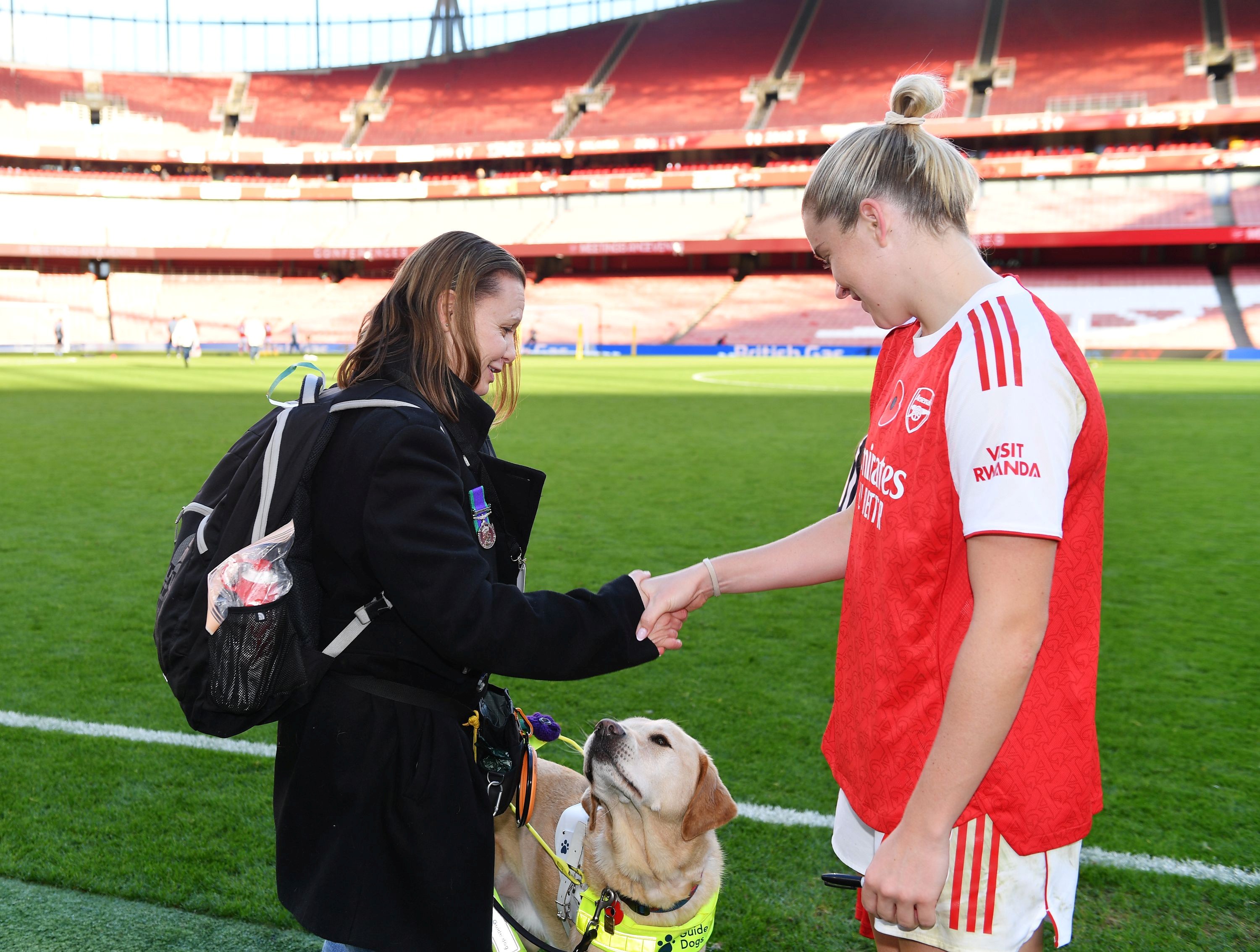 Blind veteran Kelly and her guide dog Archie are stood at the edge of the pitch at the Emirates Stadium. Archie looks up at Kelly and she shakes hands with footballer Alessia Russo. 