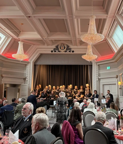 A wide shot of an event taking place. People are seated at round tables and a choir is singing on stage.