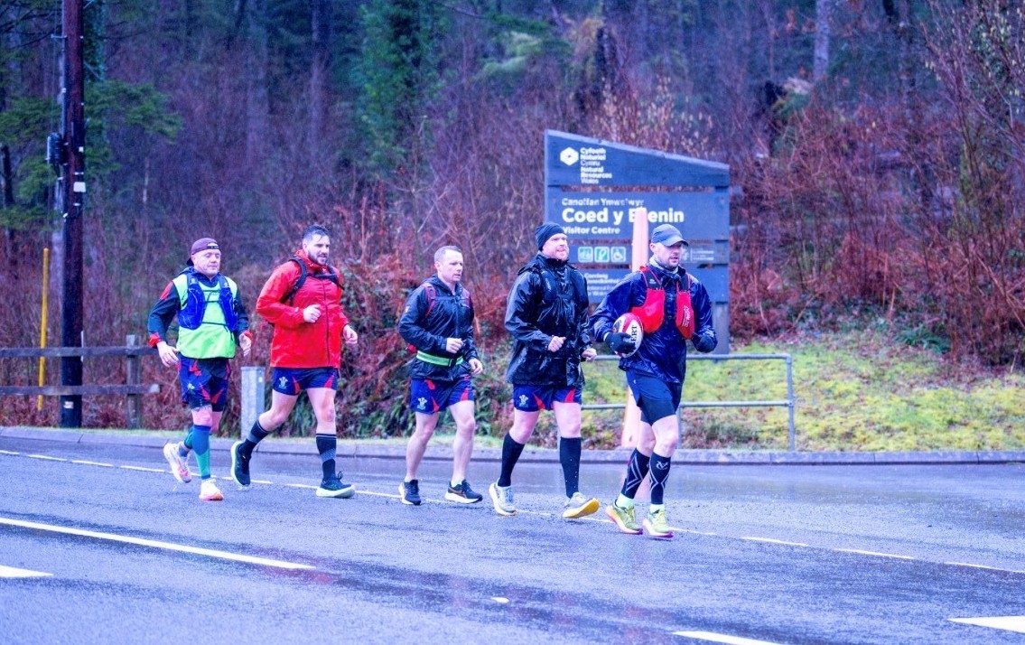 5 of the team members running in a line with the person at the front holding the rugby ball.