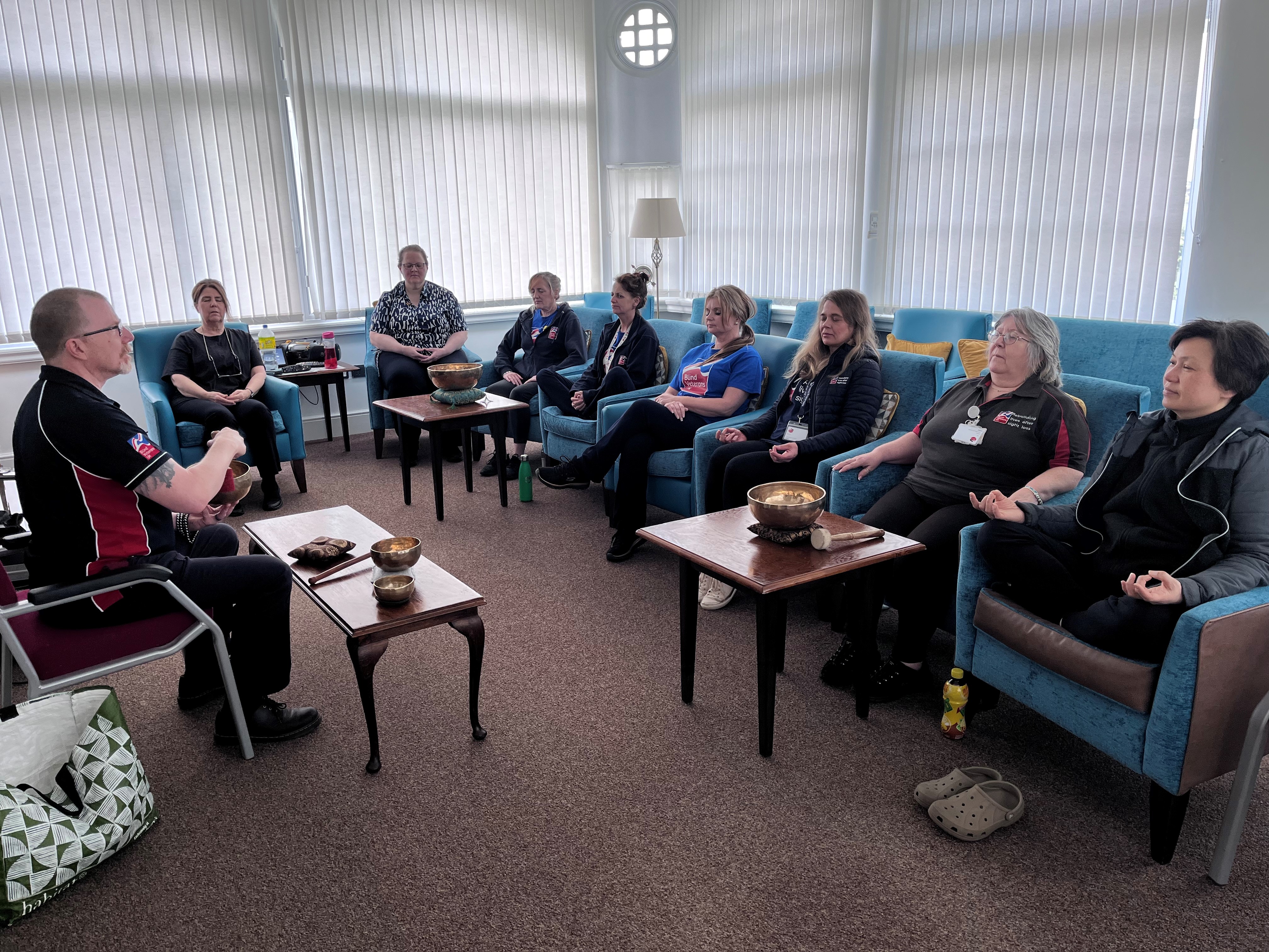 A group of people sit in chairs arranged in a circle with metal bowls placed on small tables in front of a man sat at the front of the group.