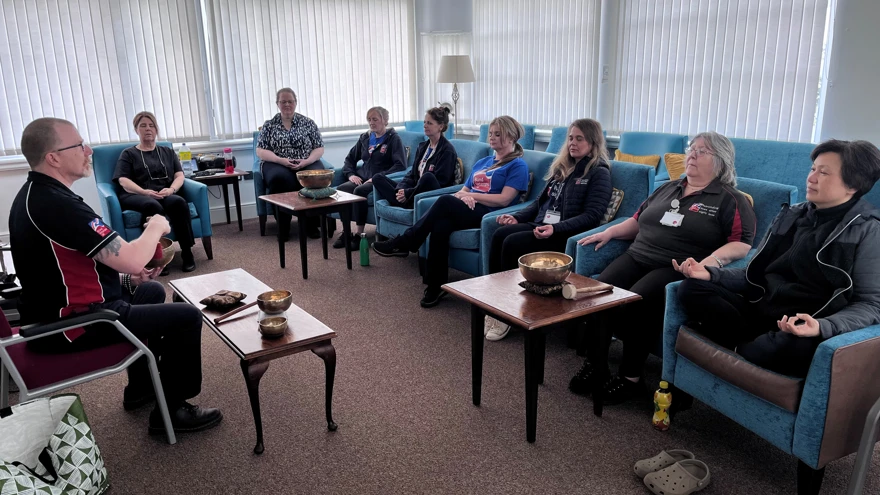 A group of people sit in chairs arranged in a circle with metal bowls placed on small tables in front of a man sat at the front of the group.