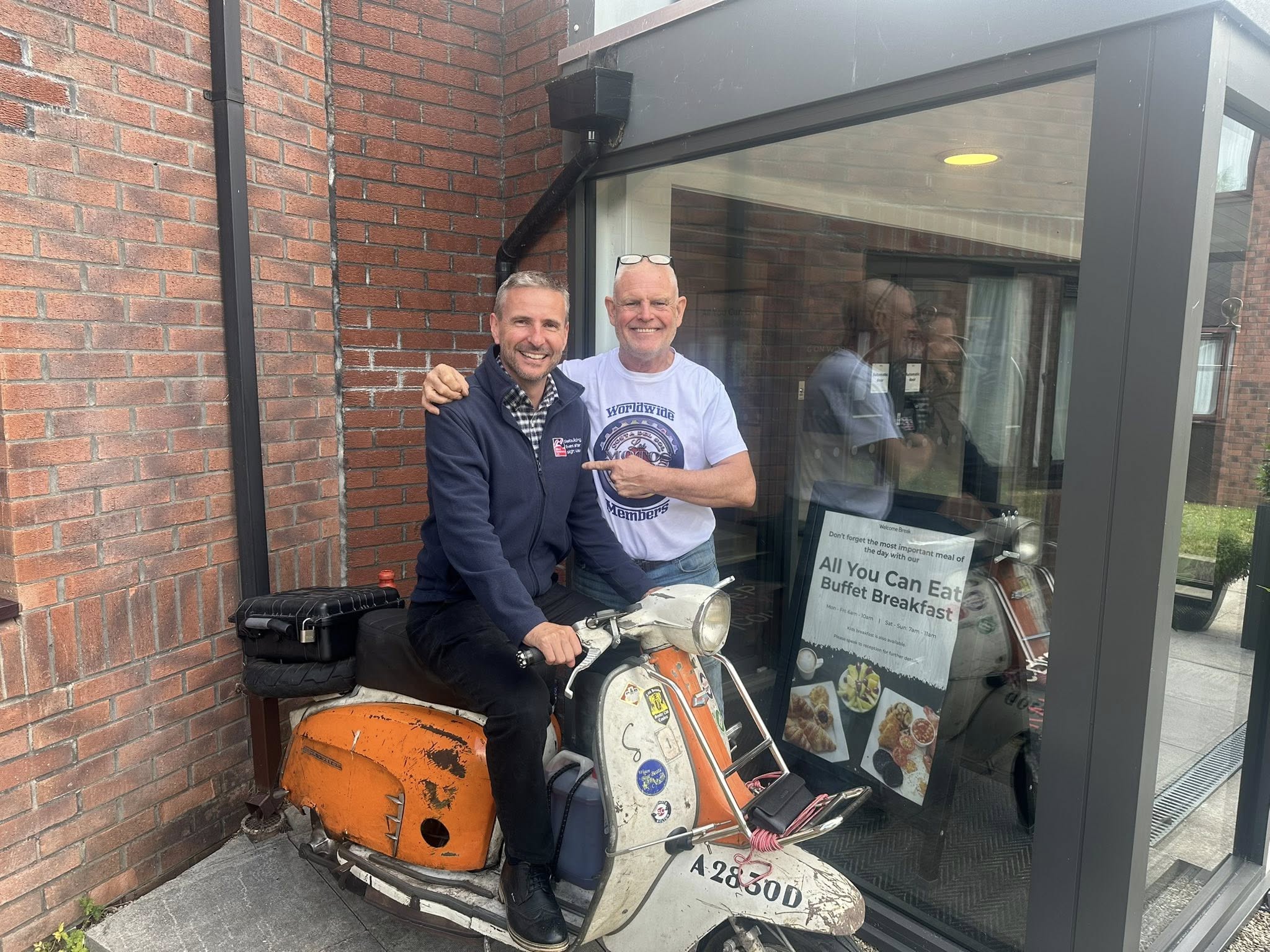 Staff member Colin wearing a Blind Veterans UK fleece sat on Paul's scooter with Paul stood beside him