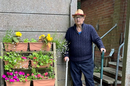 Lawrence is holding a walking stick in one hand and a garden fork in the other next to six pots with yellow and pink flowers