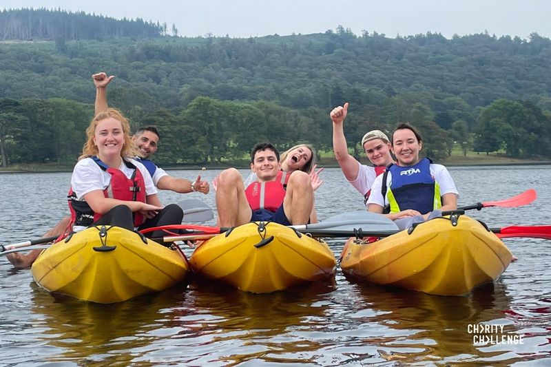 A group of event participants in 2 man kayaks on the lake, with tree covered hills in the background. The group are all smiling at the camera.  Some are holding their arms up while signalling a thumbs up.