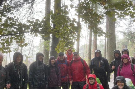 A group picture of the walkers paused under some trees. The ground is wet and they are soaked. Fully kitted out in waterproof coats and waterproof trousers.